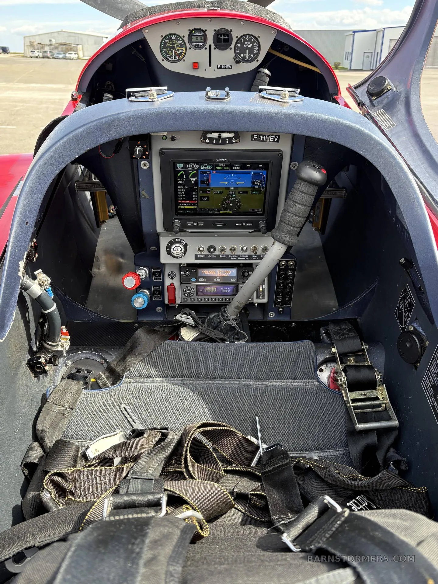 The interior cockpit of a single-seat aircraft, showing the instrument panel with a large display, control stick, and safety harnesses.