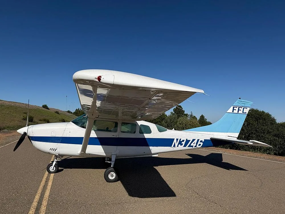 A white and blue high-wing single-engine aircraft N3746 is parked on an asphalt strip with rolling hills in the background under a clear blue sky.