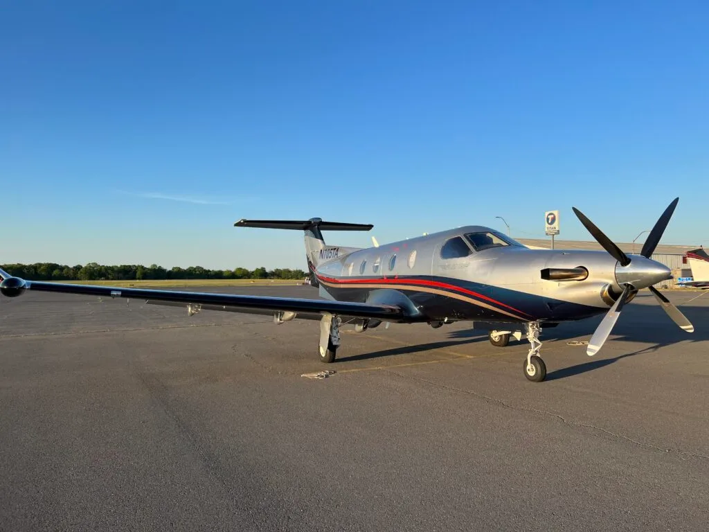 A silver and black Pilatus PC-12 turboprop aircraft is parked on an airport tarmac at sunset.