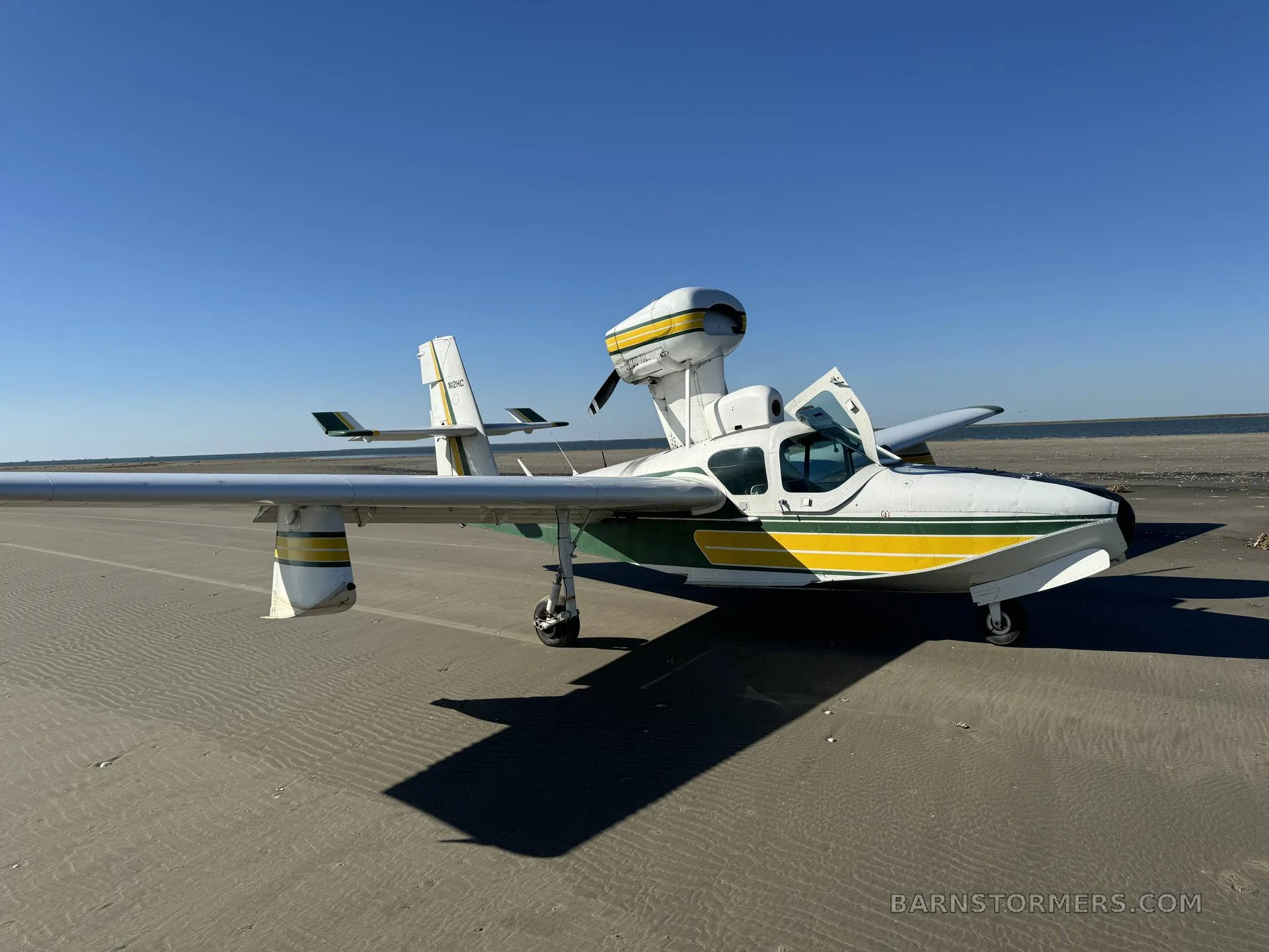 A white and green seaplane with its doors open is parked on a sandy beach under a clear blue sky.