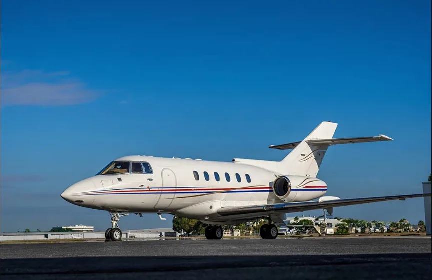 A white business jet with red and blue stripes on the fuselage is parked on an airfield under a clear blue sky.