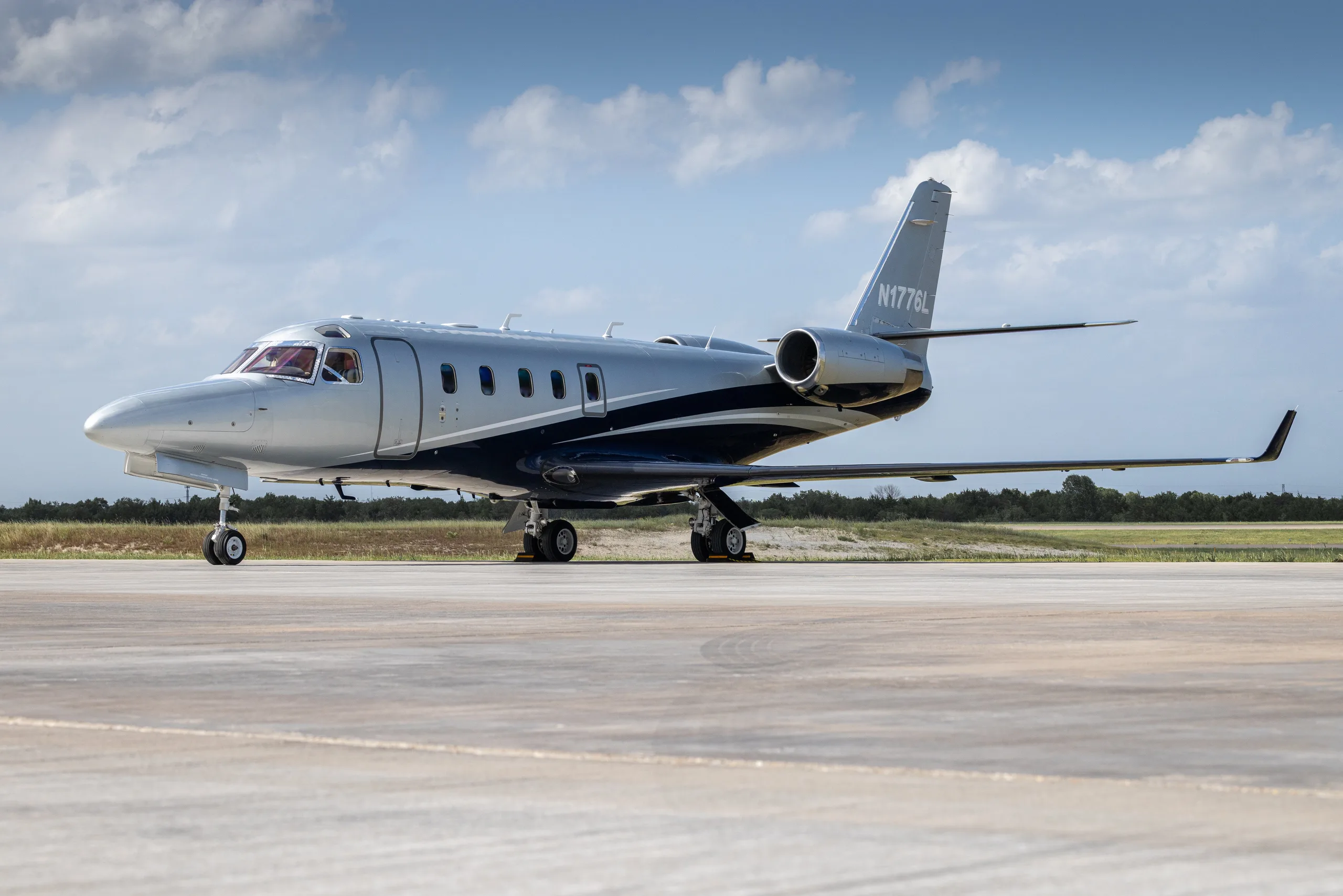 A Learjet 60 in a grey and dark blue paint scheme, parked on an airport tarmac with a bright sky.