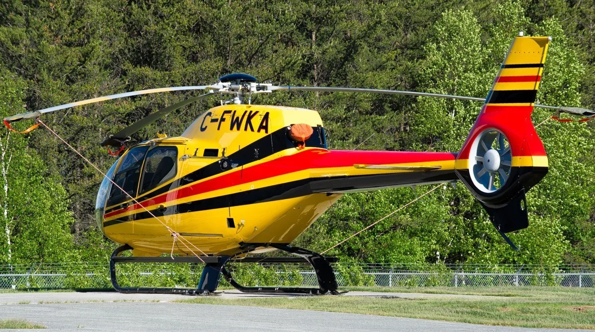 A yellow helicopter with red and black stripes, registration C-FWKA, is parked on a paved surface with trees in the background.