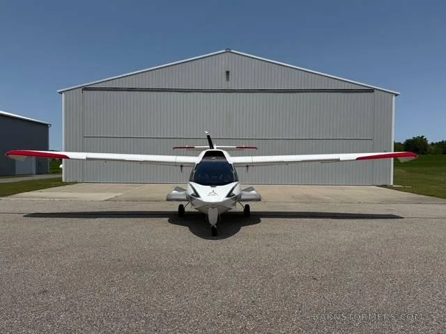 A white and silver ICON A5 amphibious aircraft is viewed head-on, parked in front of a large hangar.