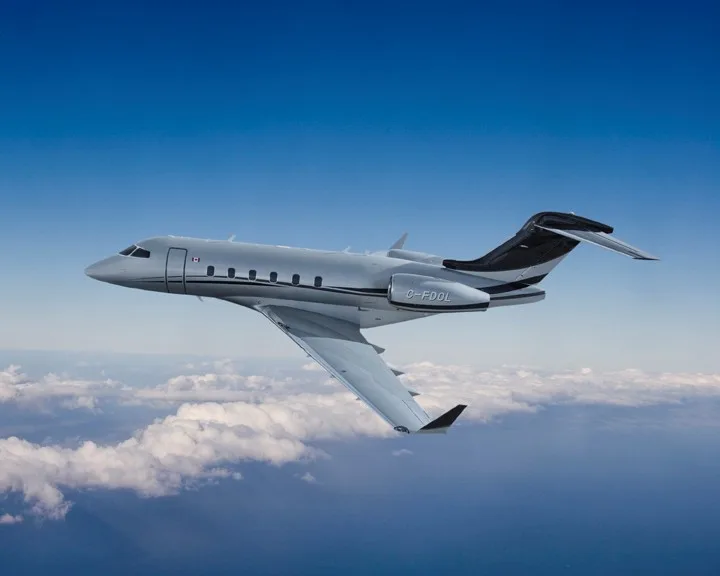 A silver and black Bombardier Challenger 605 business jet with registration C-FODL flies above clouds against a blue sky.