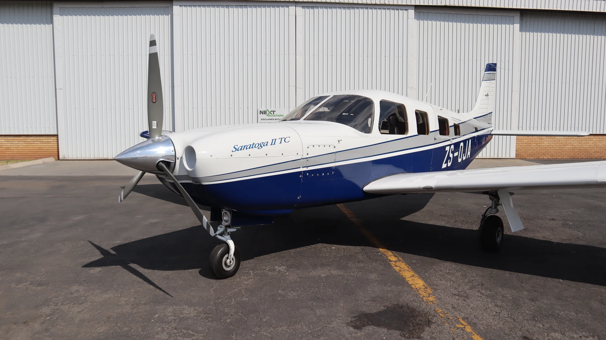 A white and blue Piper Saratoga II TC aircraft is parked on an asphalt in front of a white hangar.