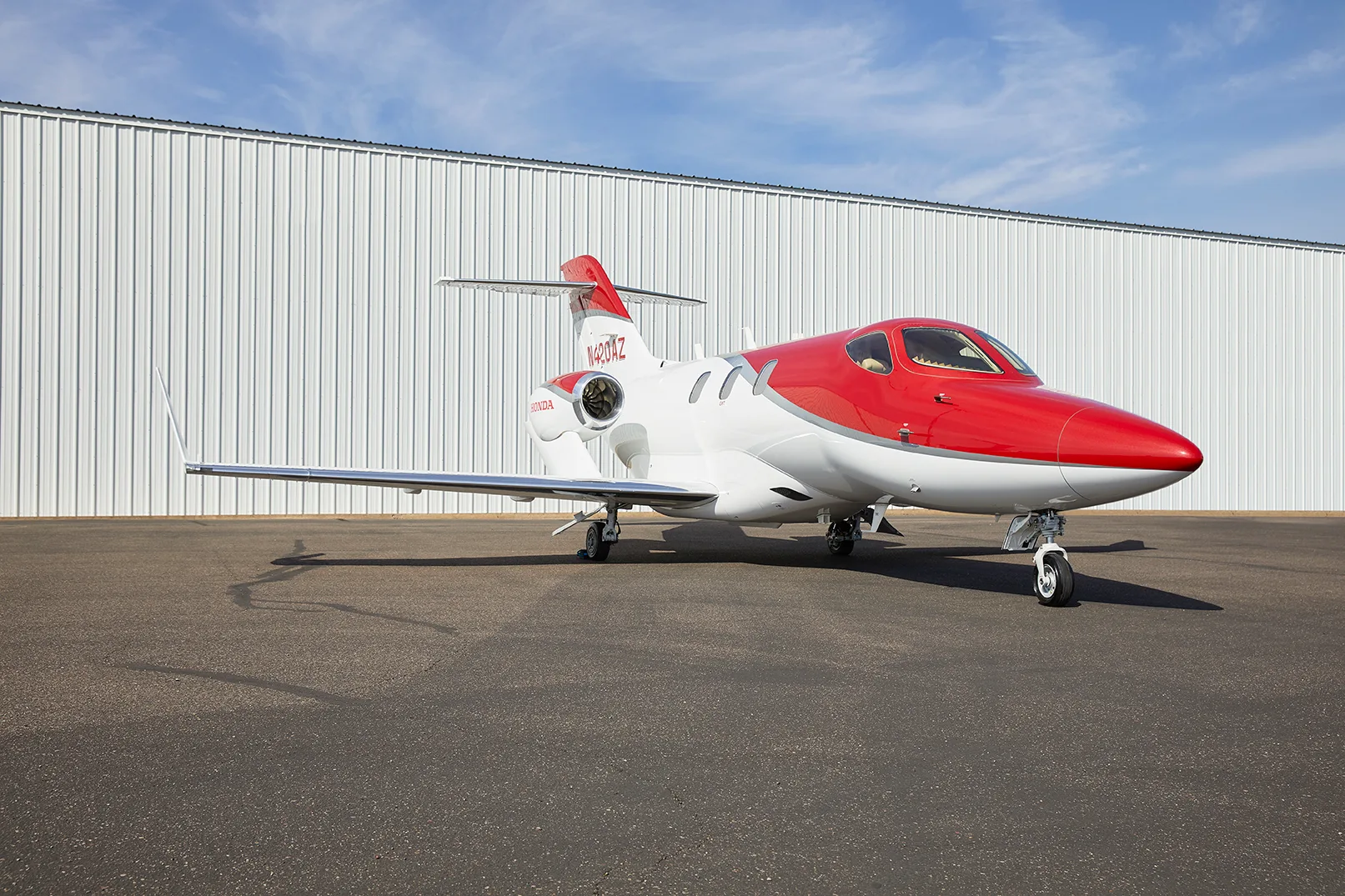 A red and white HondaJet HA-420 aircraft, registration N420AZ, is parked on an asphalt tarmac in front of a white hangar.