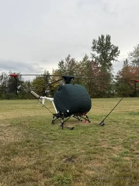 A helicopter with a dark green cover over its main body sits on a grassy field with trees in the background under an overcast sky.