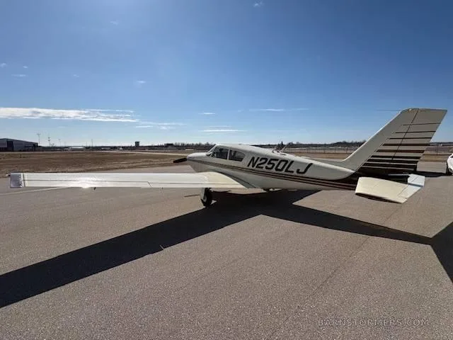 A cream-colored Piper Comanche 250 aircraft with brown stripes, seen from the side, parked on an airfield under a blue sky.