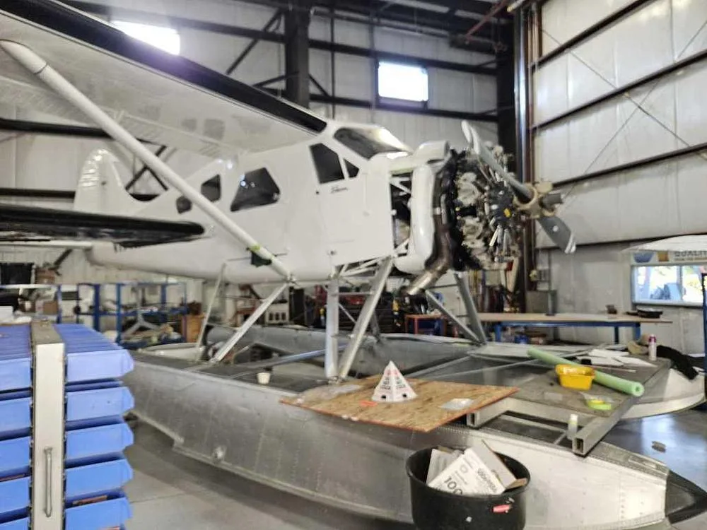 A white floatplane with its engine cowling removed, undergoing maintenance in a hangar.