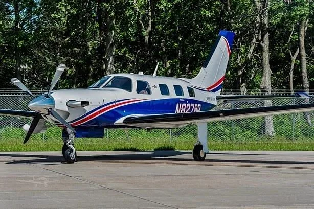 A white and blue single-engine turboprop aircraft, likely a Piper M600 with registration N827BP, parked on an asphalt ramp with trees in the background.