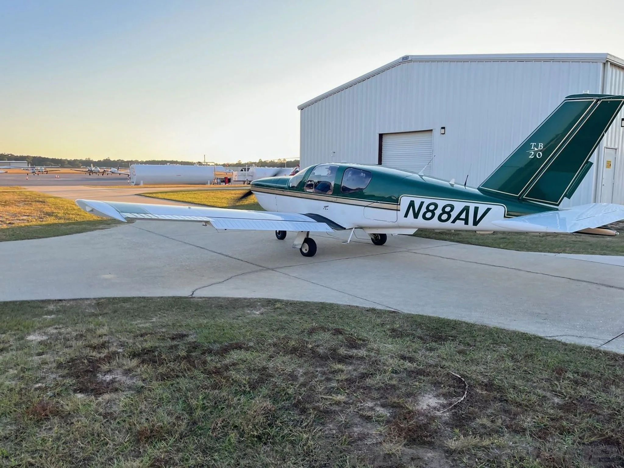 A green and white single-engine aircraft (N88AV) parked on a tarmac next to a hangar at sunset.