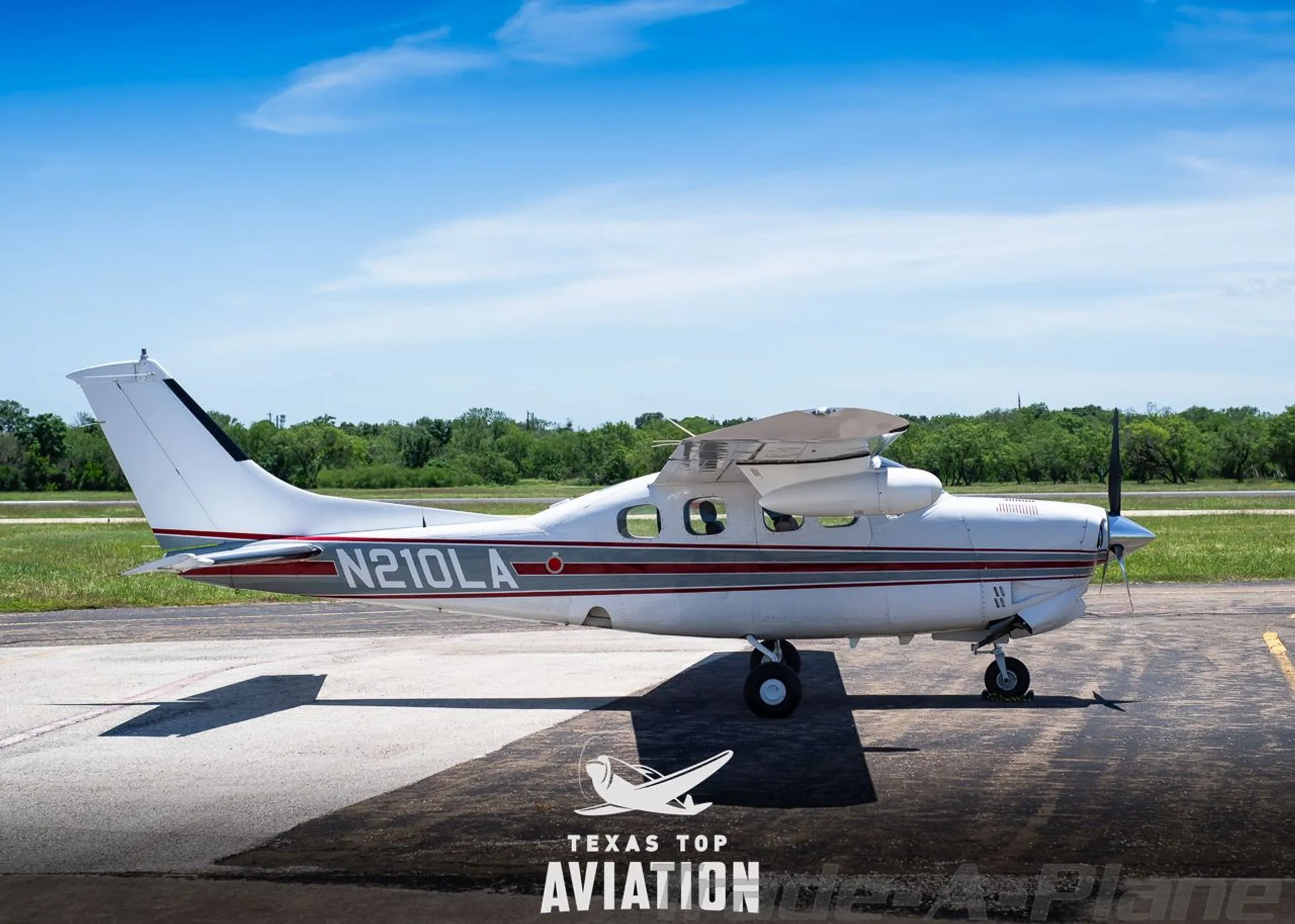 A white Cessna 210 Centurion (N210LA) with red and gray stripes parked on an airfield, viewed from the side.