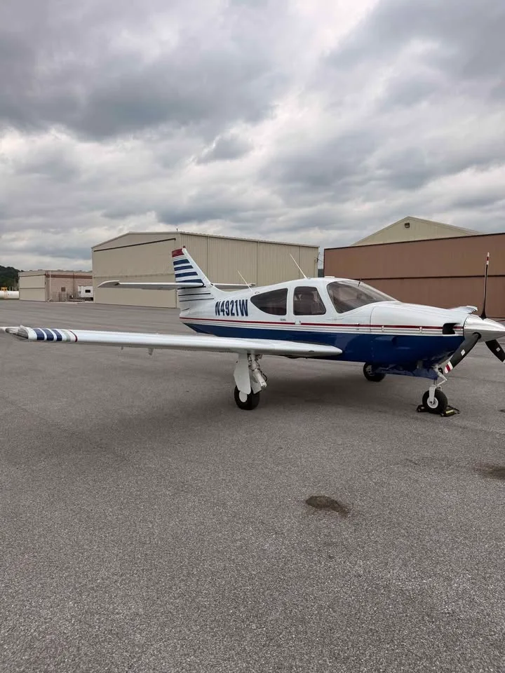 A side view of a white and blue single-engine aircraft with registration N4921W parked on the tarmac in front of hangars.