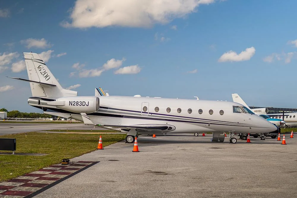 A white Gulfstream G200 business jet, tail number N283DJ, is parked on an airport tarmac under a partly cloudy sky.