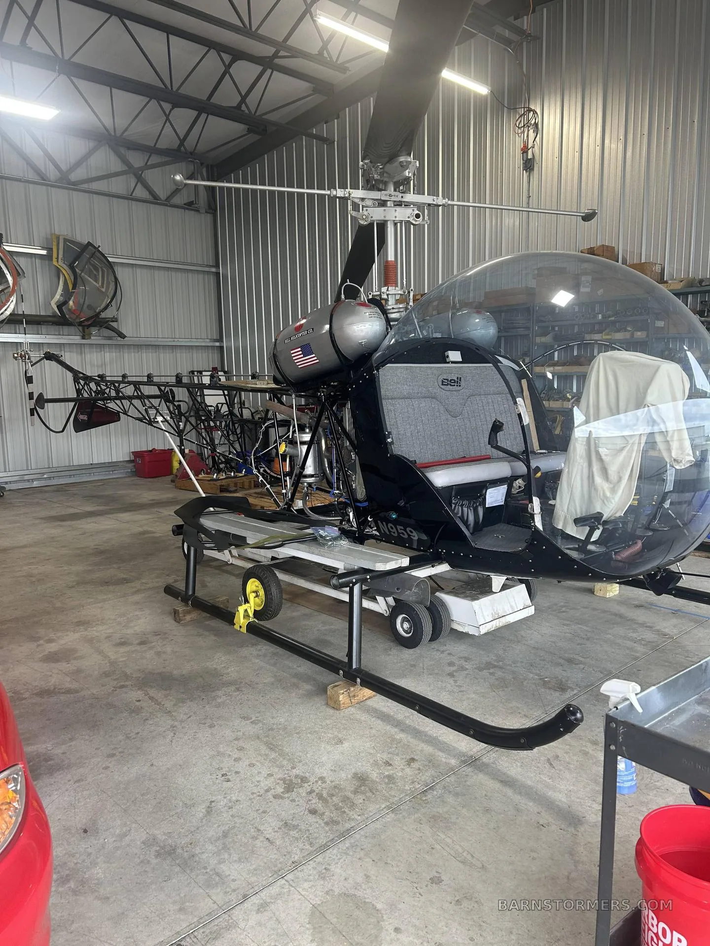 A black Bell 47 helicopter, partially assembled, sits on a stand in a hangar.