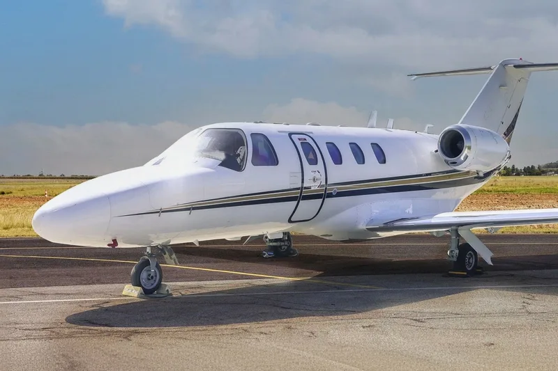 A white business jet with black and gold stripes is parked on a tarmac with its passenger door open, under a cloudy sky.