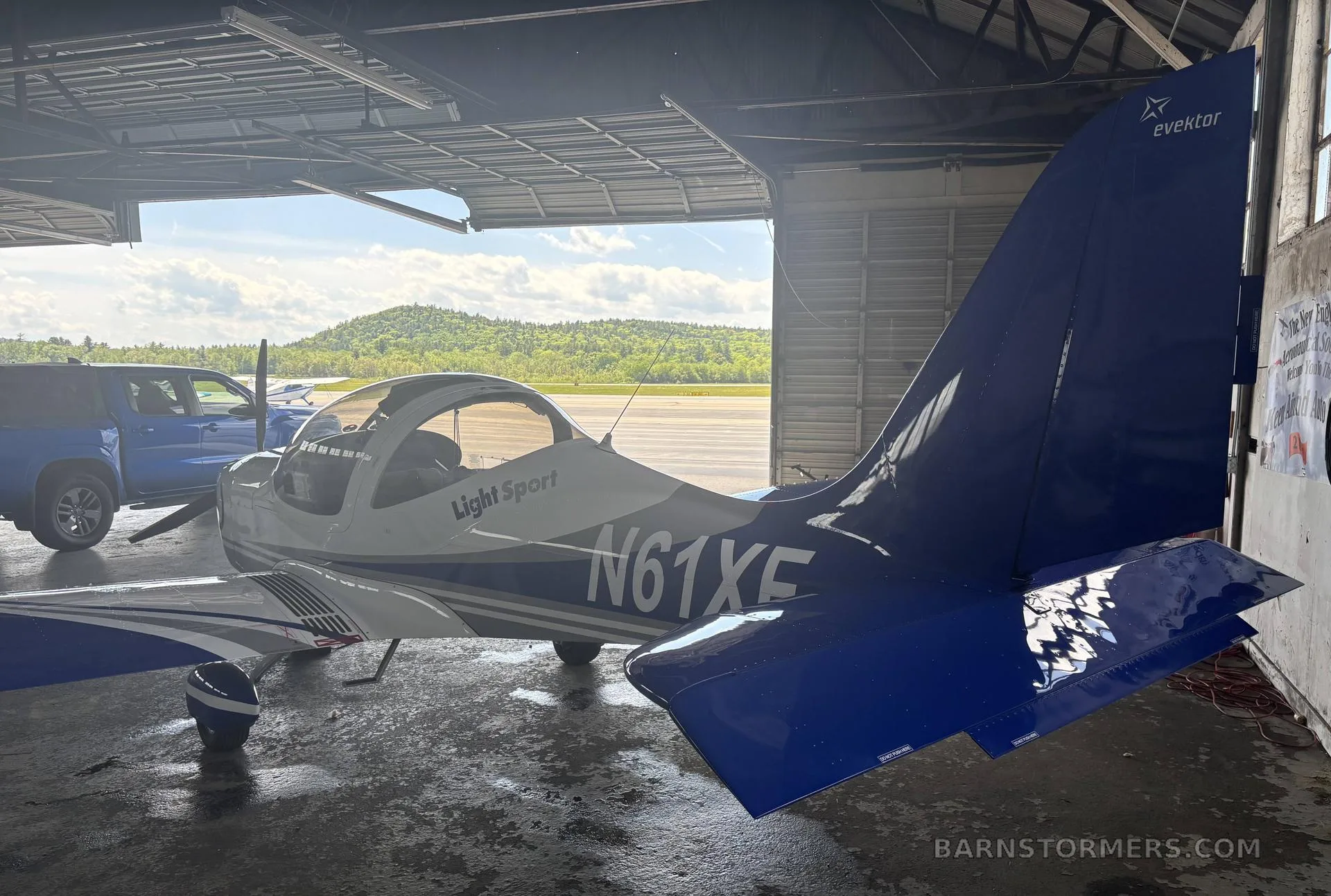 The tail and rear fuselage of a white and blue Evektor Harmony Light Sport aircraft (N61XF) are seen inside a hangar, looking out to an airfield.