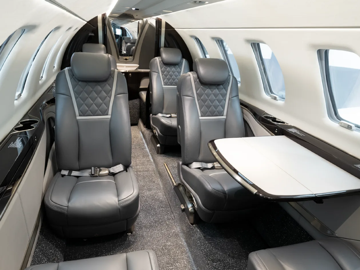 The luxurious interior cabin of a business jet, featuring grey leather seats with diamond stitching and pull-out tables.