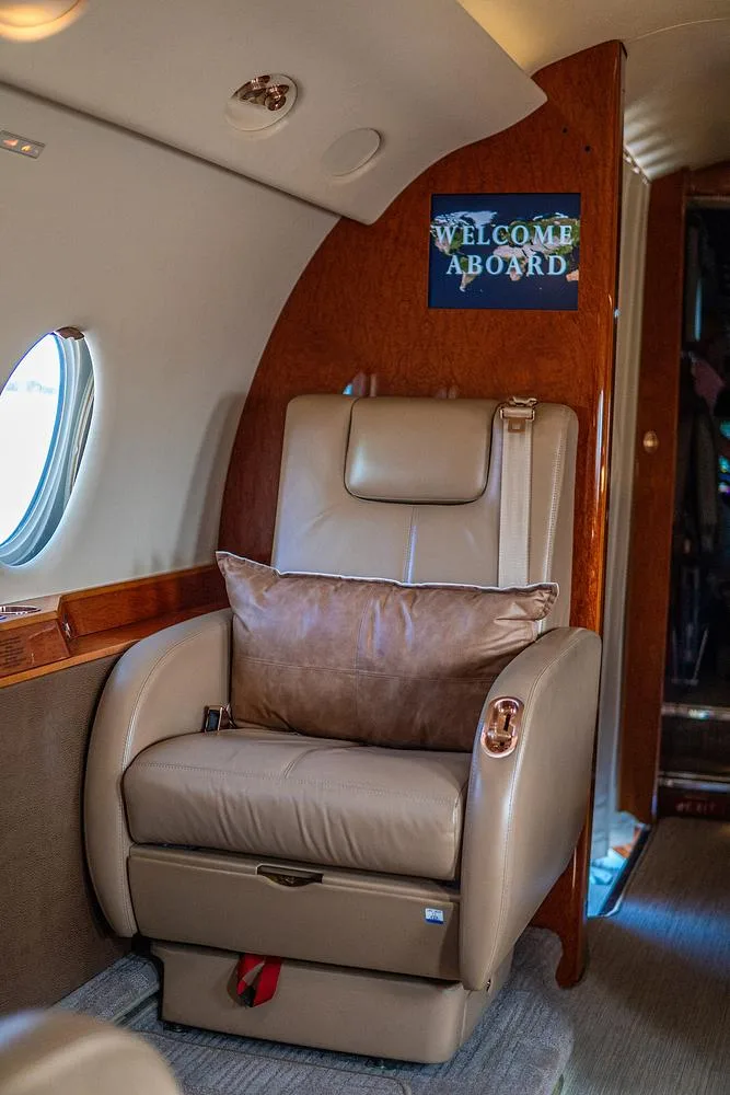 A tan leather seat with a brown pillow is visible in the private jet cabin, next to a "WELCOME ABOARD" sign.
