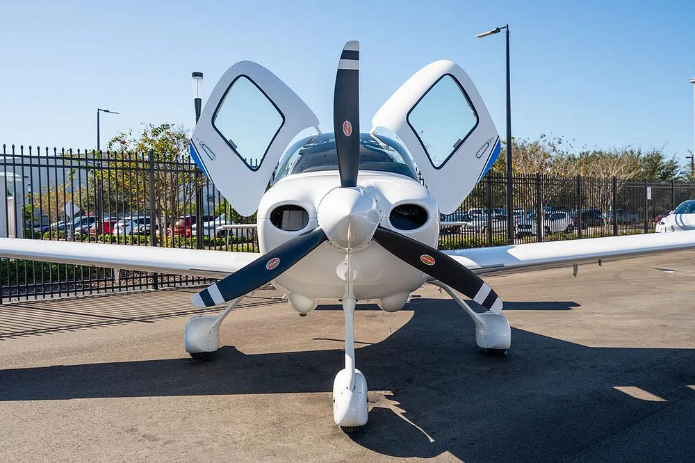 Front view of a white Cirrus SR22 aircraft with blue and black stripes, registration N323HW, showing its propeller and open gull-wing doors.
