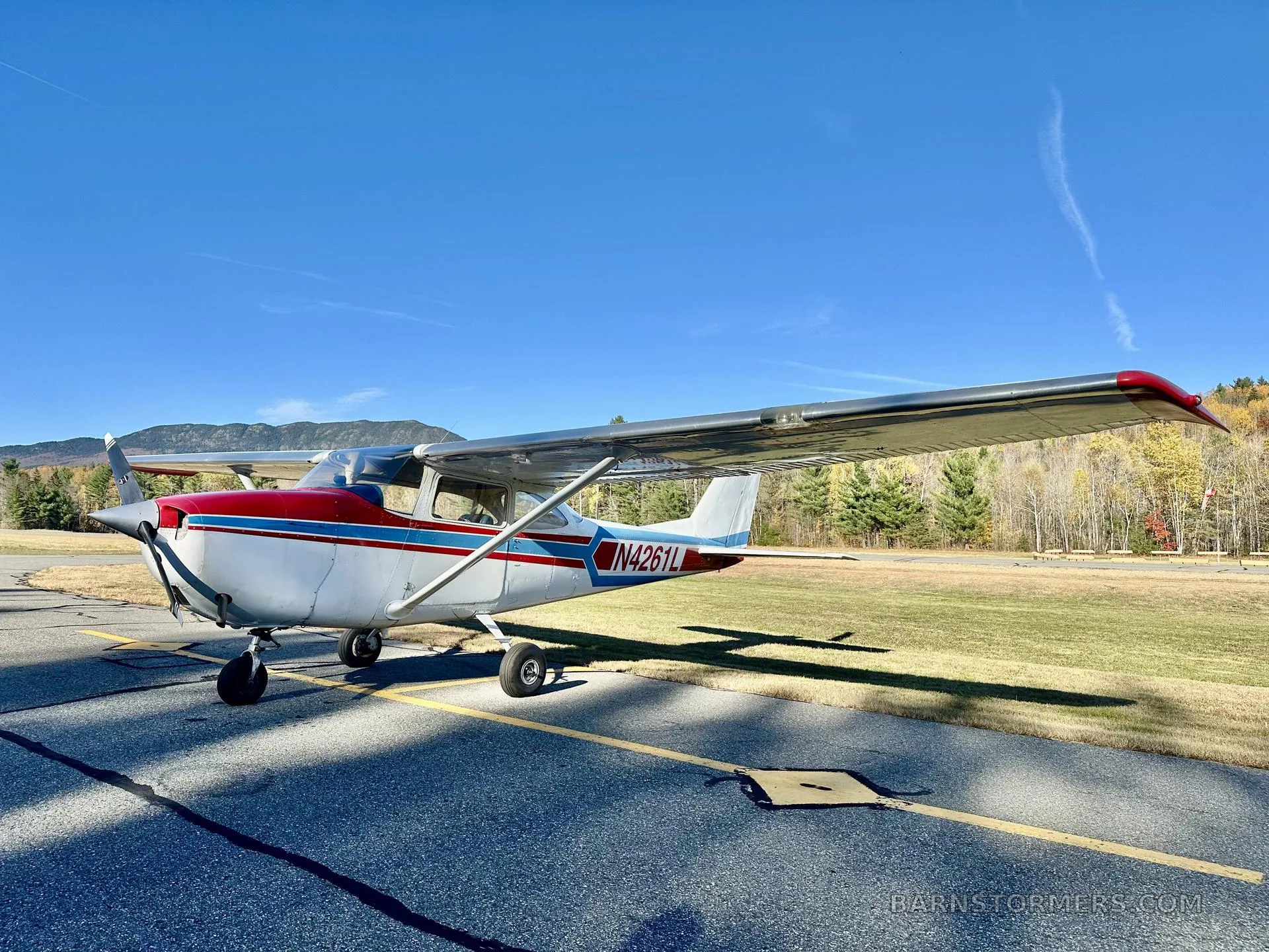 A white single-engine propeller aircraft with red and blue stripes, viewed from the front-left, parked on an asphalt ramp with autumn trees and mountains under a clear sky.