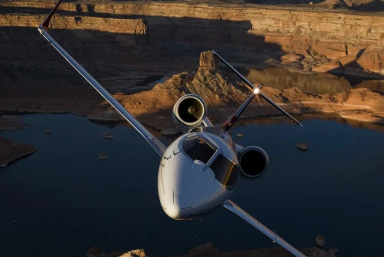 A white business jet with red accents flying over a scenic canyon and lake at sunset.