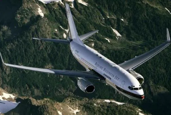 A Boeing Business Jet flies over a mountainous landscape.