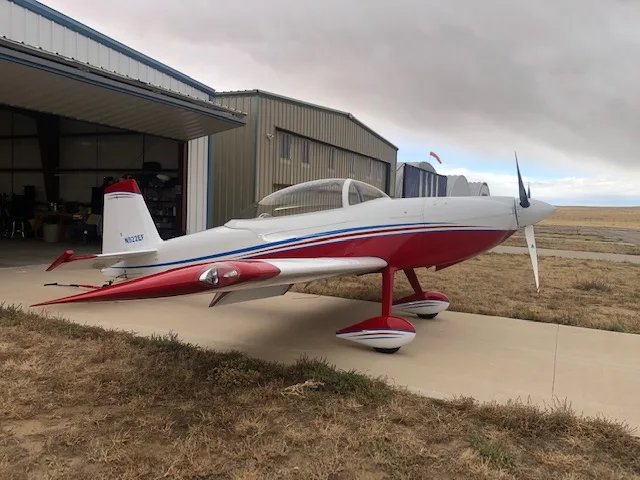 A white and red single-engine sport aircraft with blue stripes parked on an apron outside a hangar.