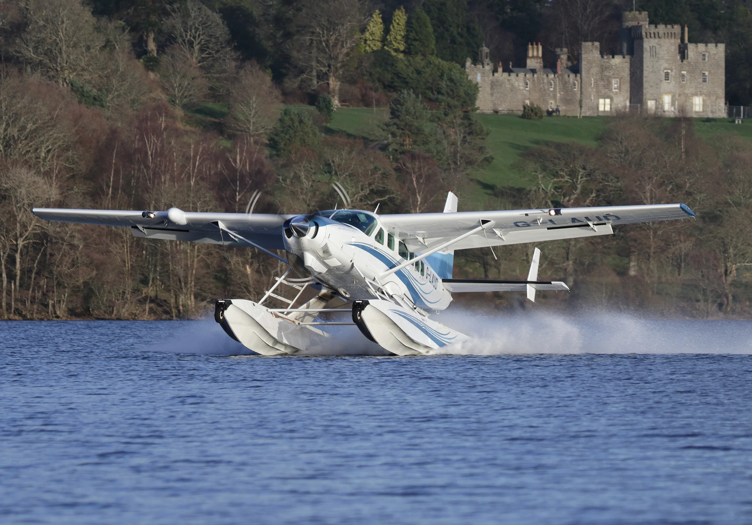 A white and blue floatplane, a Cessna Caravan, takes off from a lake, creating a spray, with a castle on a hill in the background.