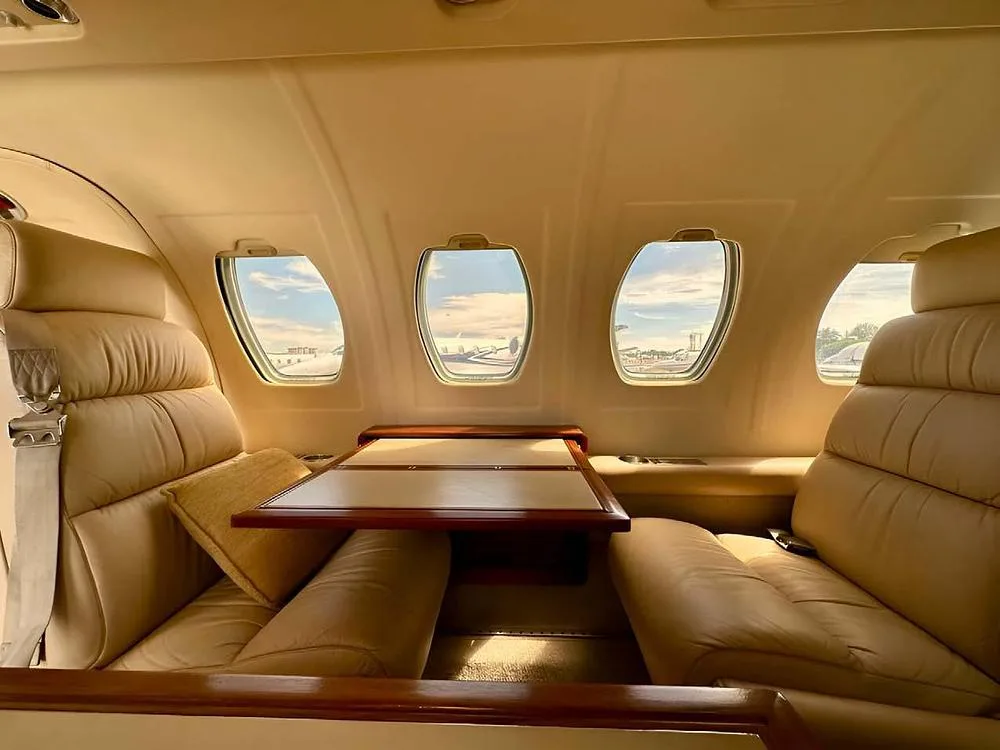 A view inside the aircraft cabin shows light tan leather seats and a wooden table next to a window.