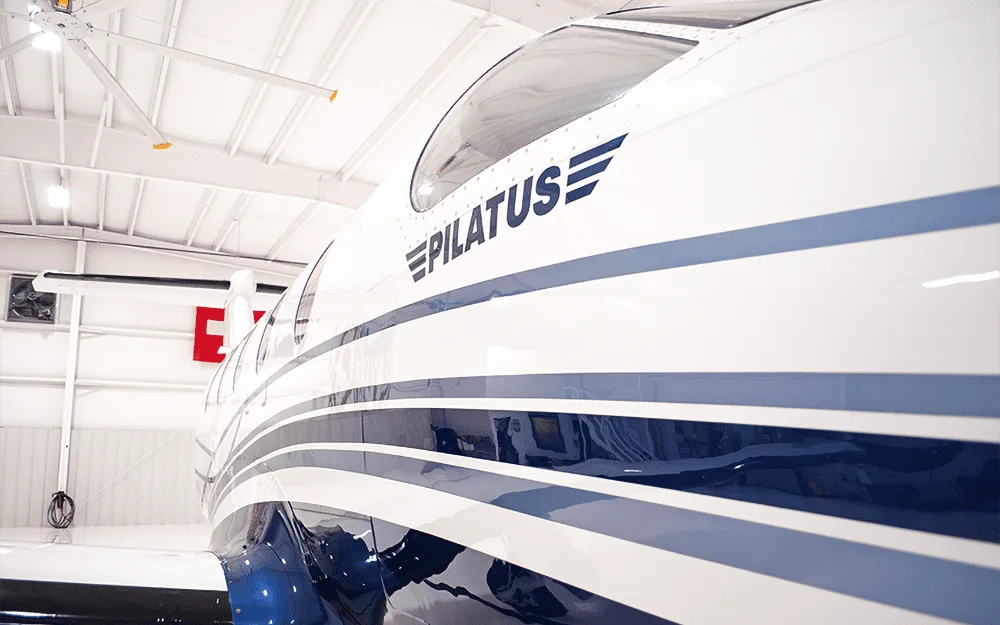 A close-up view of the white and blue fuselage of a Pilatus PC-12 aircraft, showing the "PILATUS" logo.