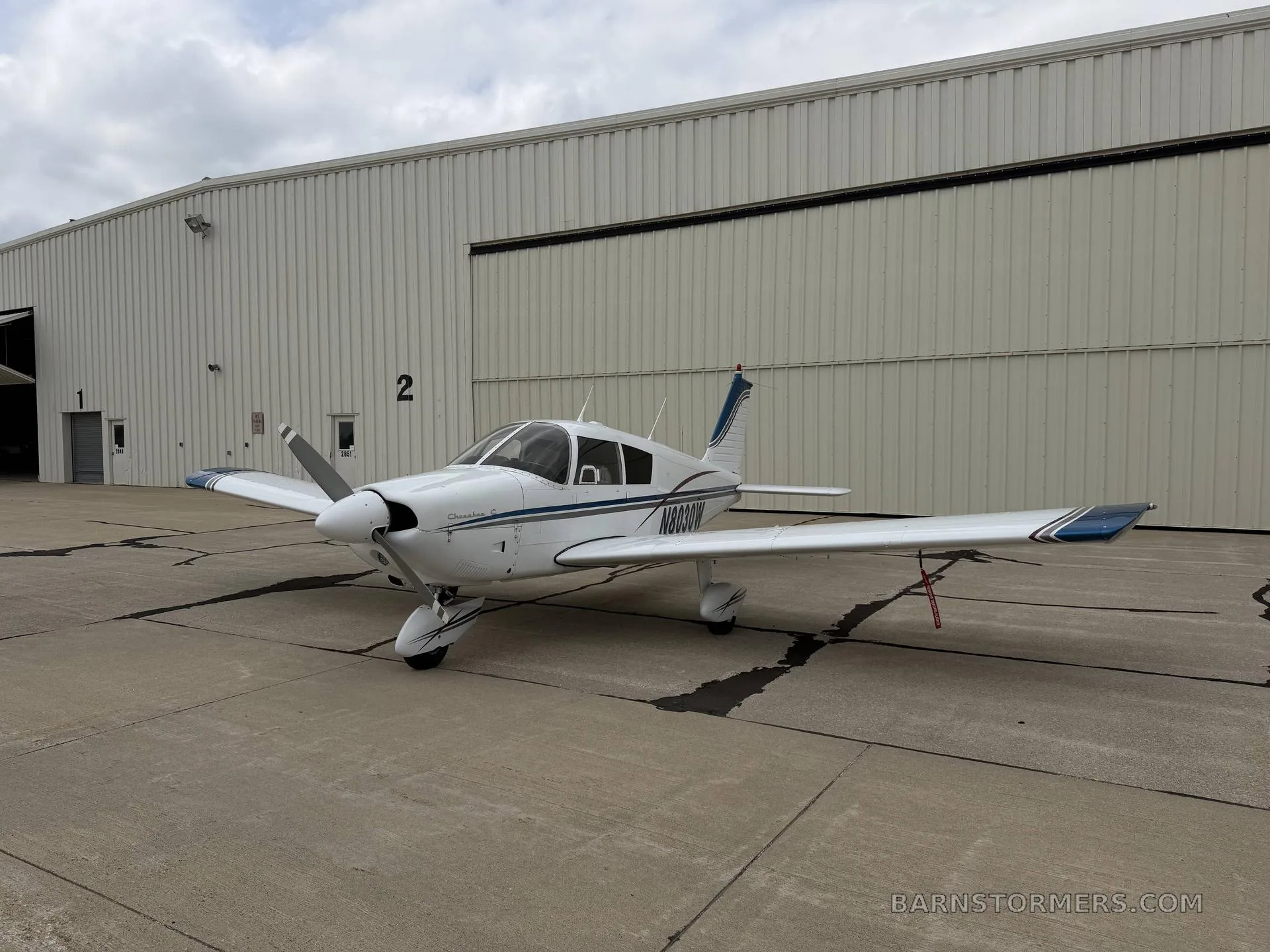 A white Piper Cherokee aircraft with blue accents, viewed from the front-left, parked on an asphalt apron in front of a hangar.