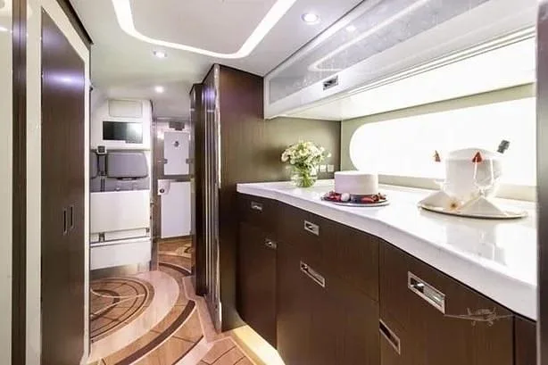 A luxurious aircraft galley with dark wood cabinetry, white countertops, and a vase of flowers, leading to a corridor with patterned flooring.