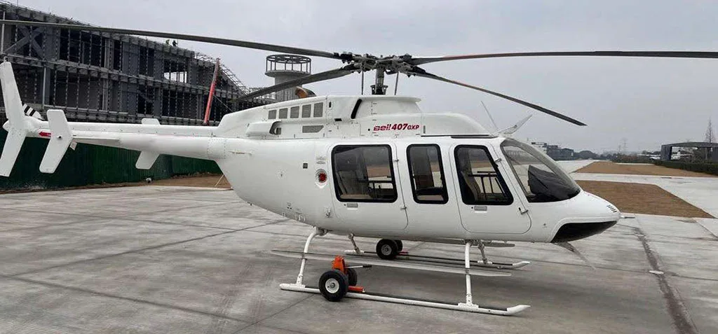 A white Bell 407GXP helicopter with landing skids and ground handling wheels, viewed from the side on a concrete pad.
