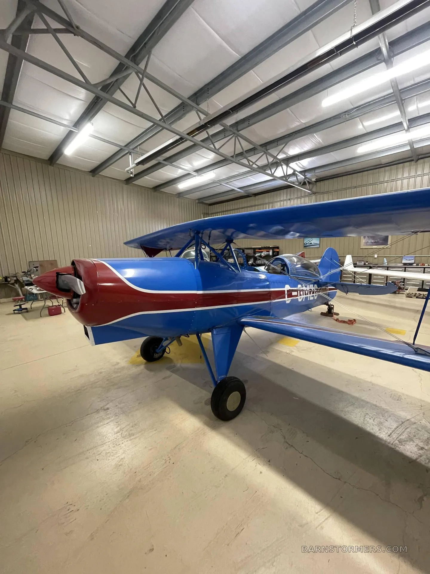 A front-quarter view of a blue and red biplane with registration C-GMZG parked in a hangar.
