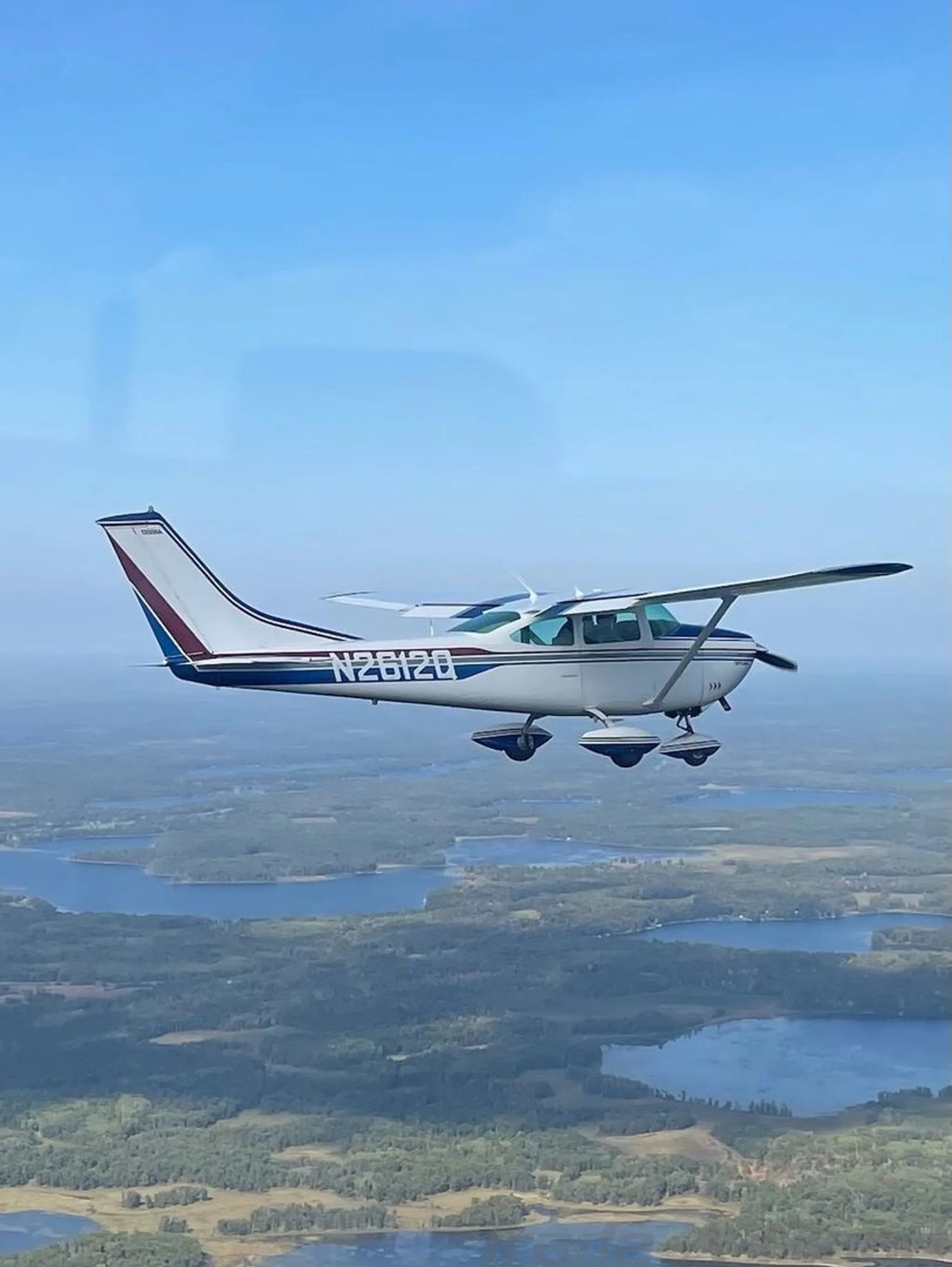 A white and blue Cessna N2612Q Skylane flying over a scenic landscape of forests and lakes.