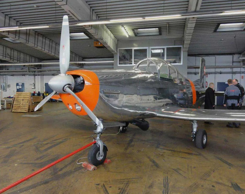 A polished silver and orange Pilatus P-3 aircraft with a propeller, parked inside a hangar with people in the background.