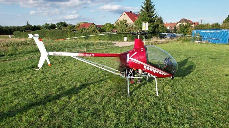 A red and white Kompress helicopter with registration OK-KHA11 is parked on a vibrant green field under a partly cloudy sky.