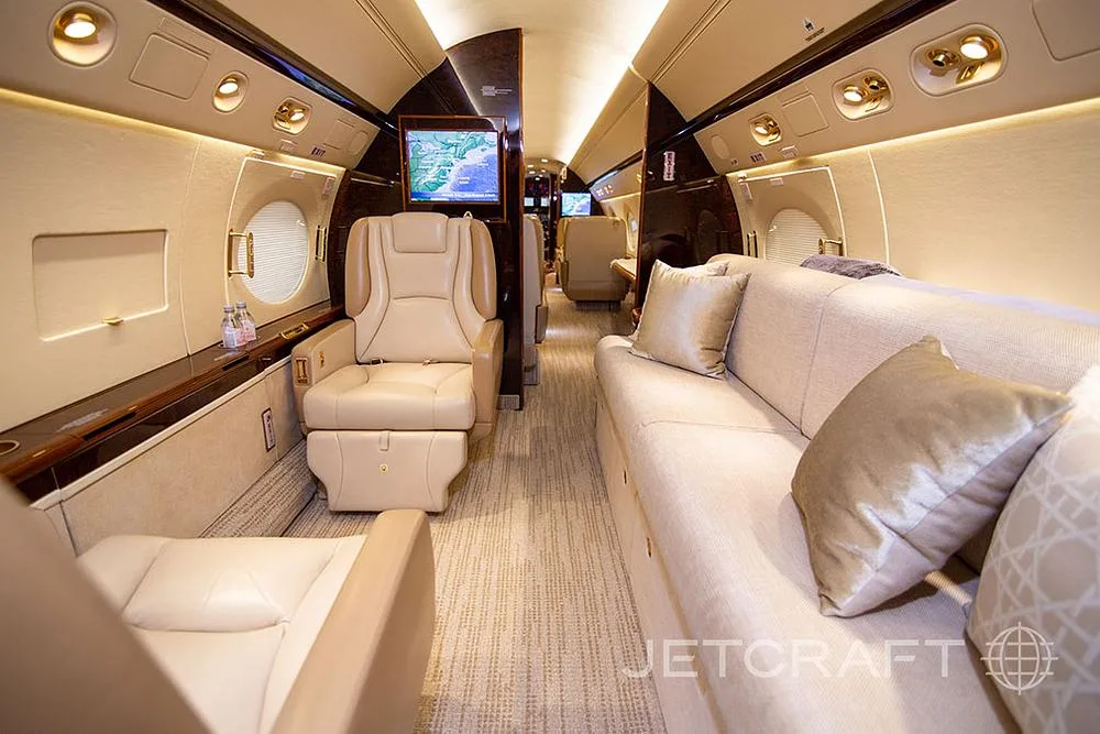 The spacious interior of a private jet, featuring a beige sofa with pillows, individual seats, and a screen.