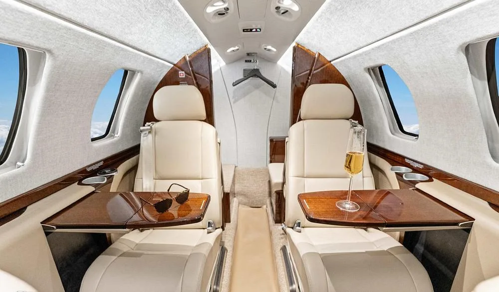 A view inside the private jet cabin showing two beige leather seats with a polished wooden table holding a glass of champagne and sunglasses.