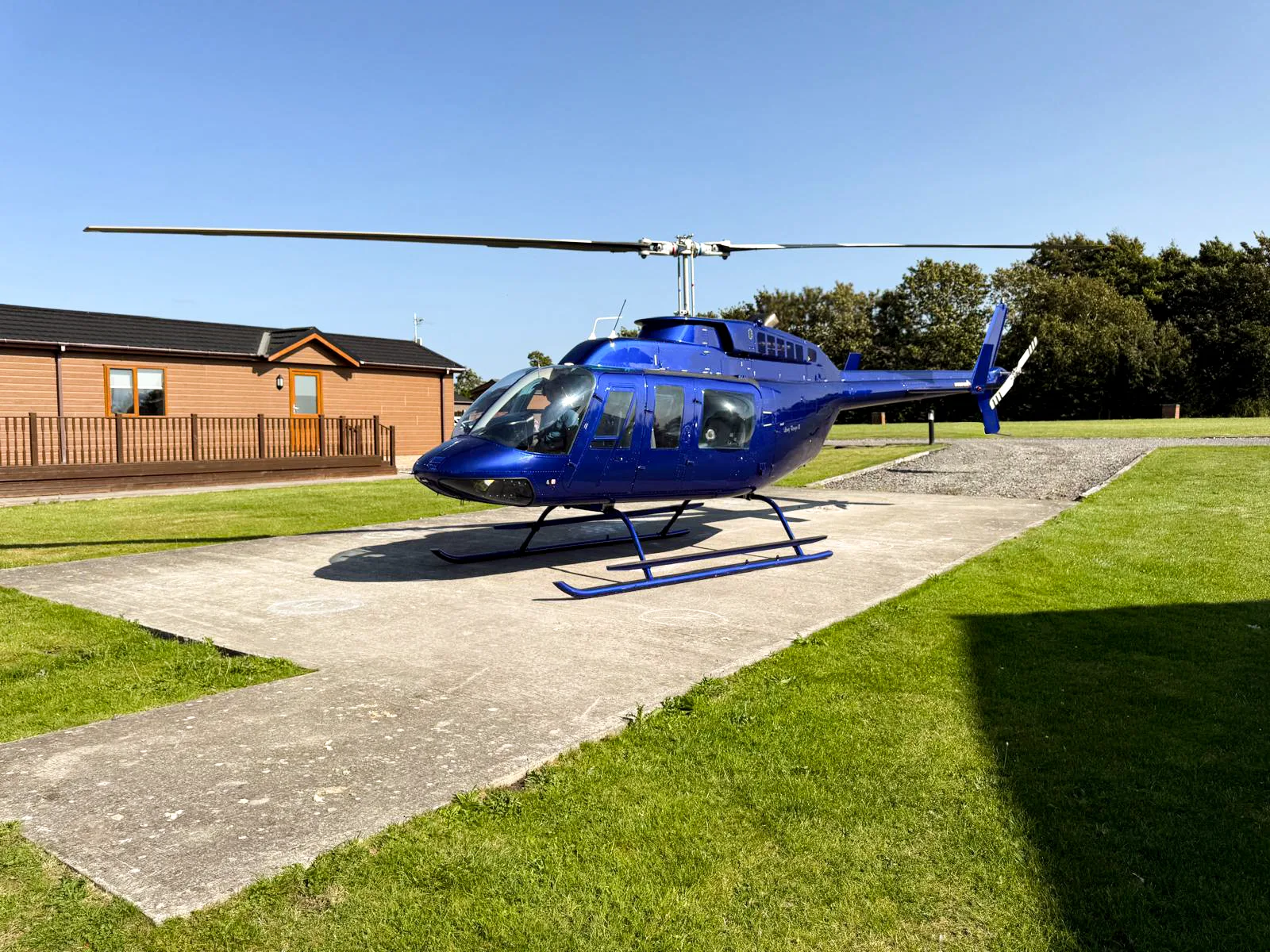 A blue helicopter with skids is parked on a concrete pad next to a wooden building under a clear blue sky.