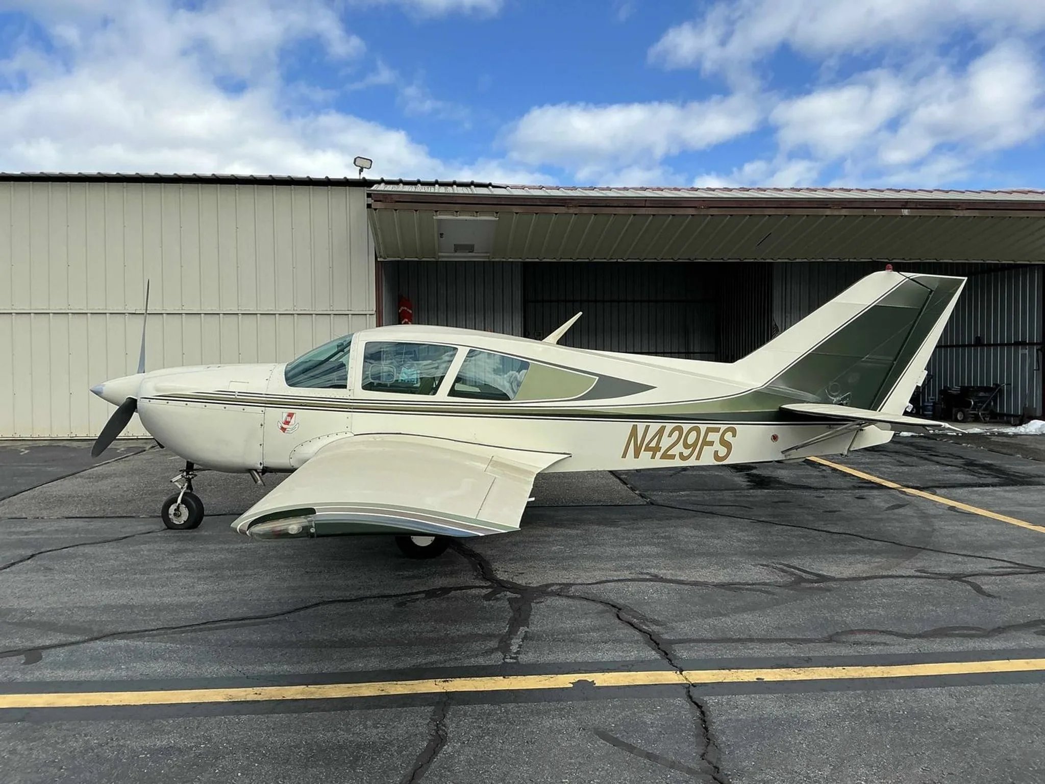 A side view of the white and green Bellanca Viking aircraft, N429FS, parked in front of an open hangar.