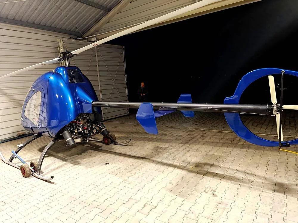 A blue single-seat helicopter with its engine exposed is parked inside a hangar at night, with a person in the background.