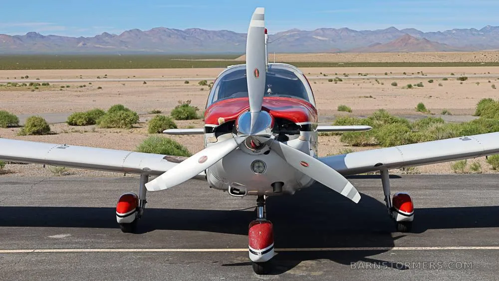 Front view of a red and white single-engine propeller aircraft on an airfield with mountains in the background.