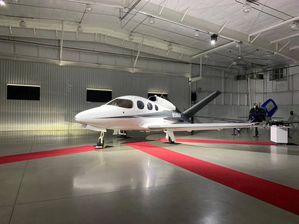 A white Cirrus Vision Jet with black and grey accents, registration N58AA, parked inside a hangar with red carpet strips.