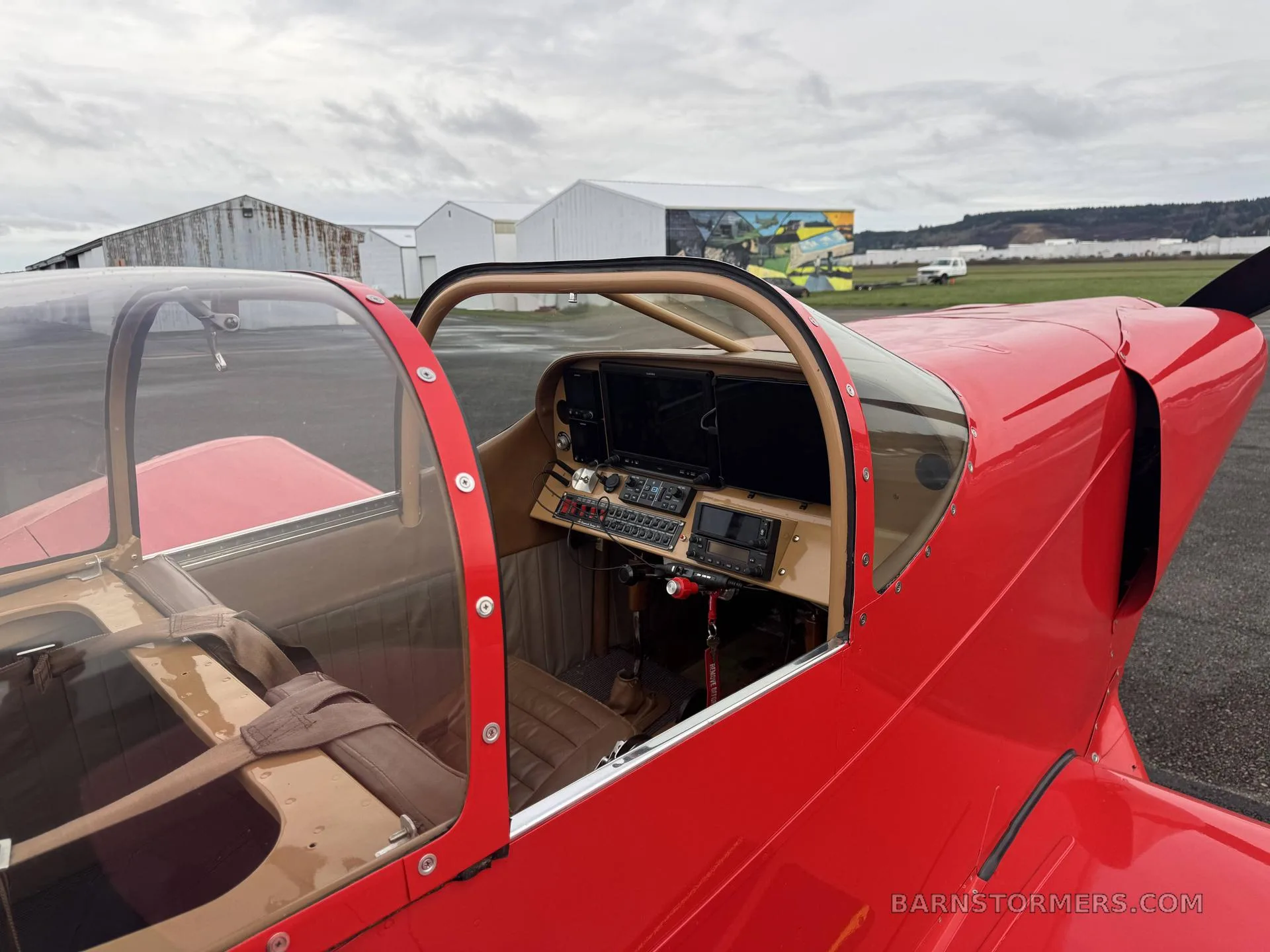 A view into the cockpit of a red experimental aircraft, showing the instrument panel with dual screens, tan interior, and an open canopy on an airfield.