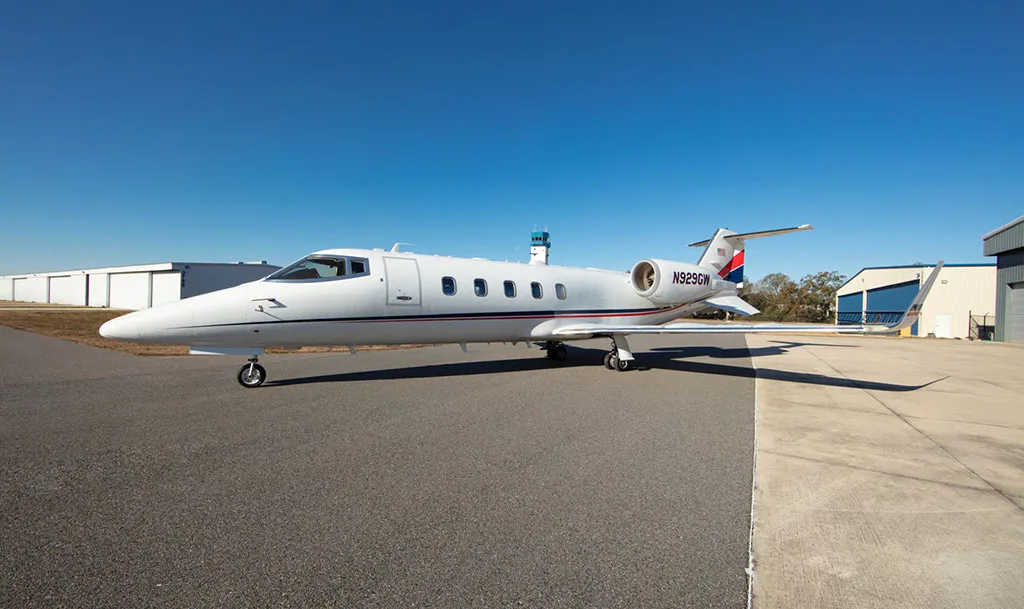 A white private jet with red and blue tail stripes, registration N929GW, parked on an airfield with hangars and a control tower in the background.