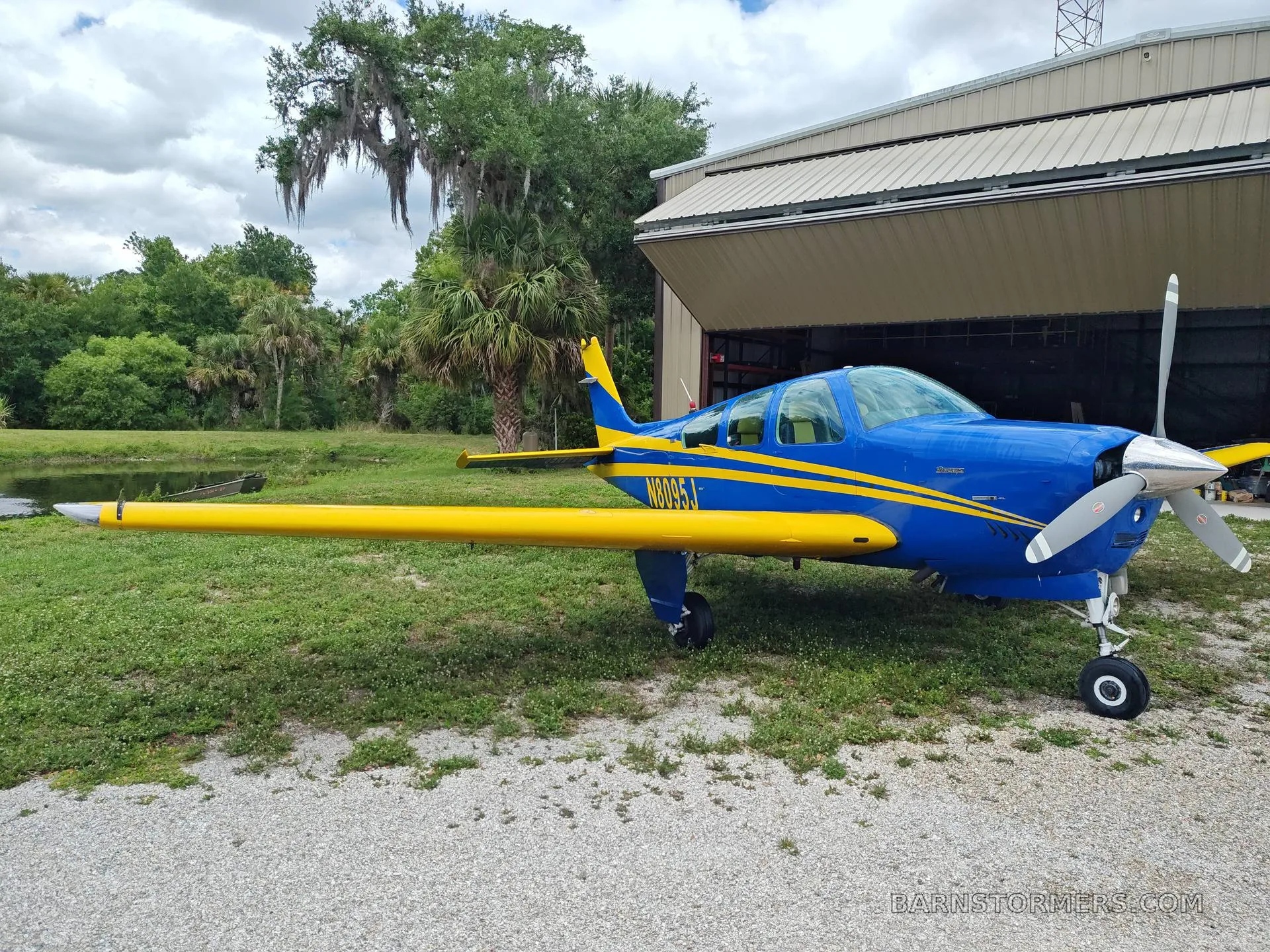 A blue and yellow single-engine aircraft is parked on a gravel path next to a hangar, with trees and a pond in the background.