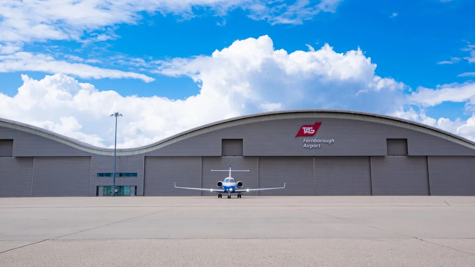 A blue and white jet sits on the tarmac in front of a hangar at Farnborough Airport.